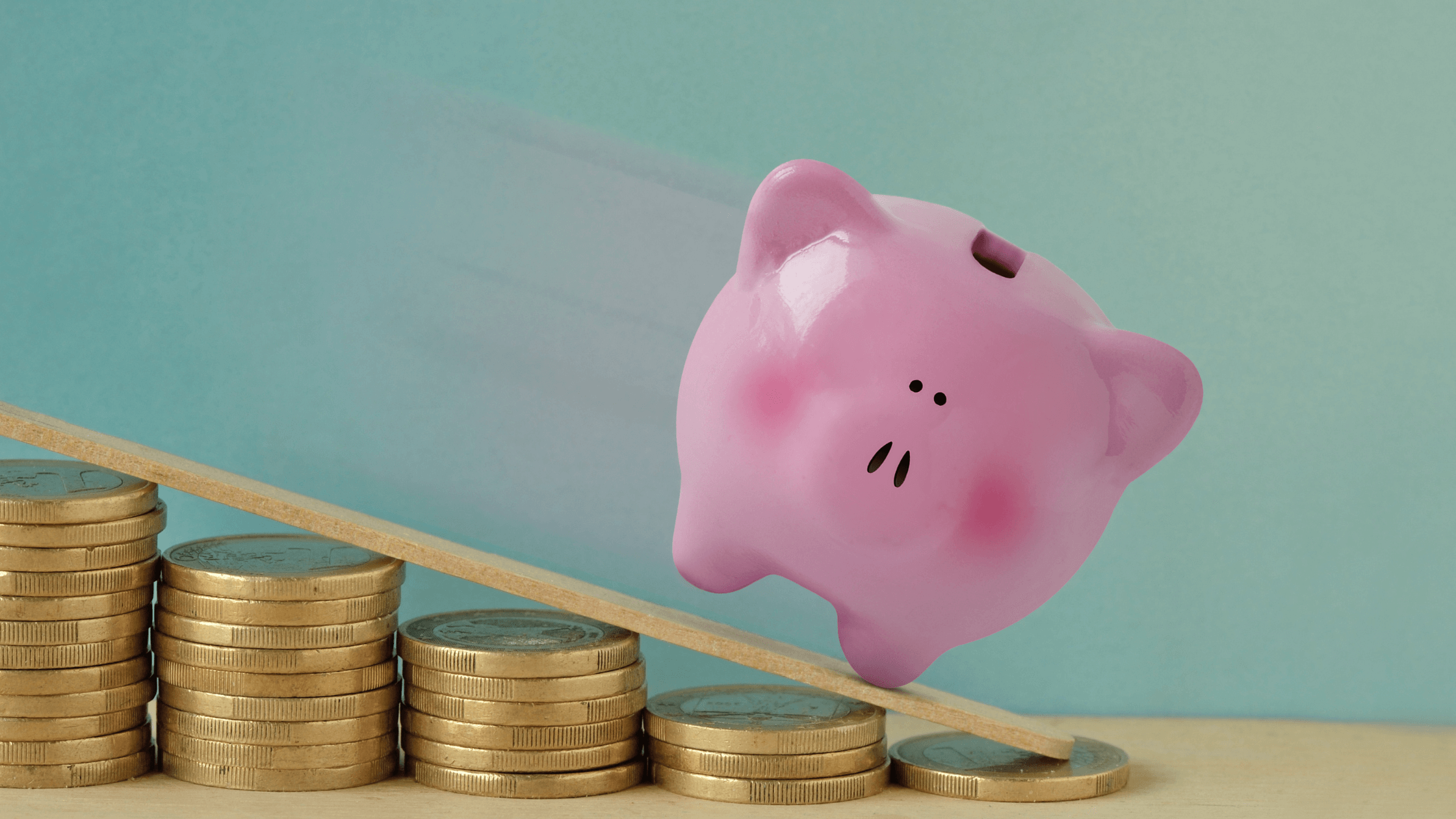 A pink piggy bank sliding down a ramp made of a wooden ruler, away from stacks of coins, representing a business losing money and the need for cash flow management.