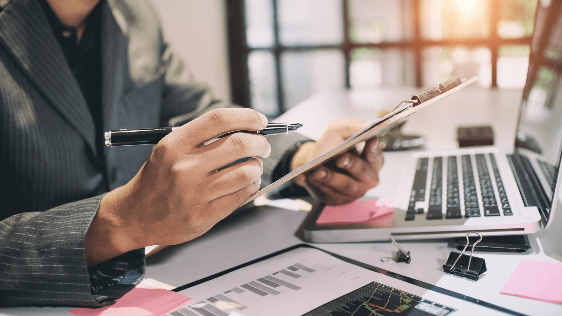 A business professional in a suit is holding a clipboard and pen, reviewing financial documents and charts on a laptop and tablet, representing proactive tax planning and financial consulting services.