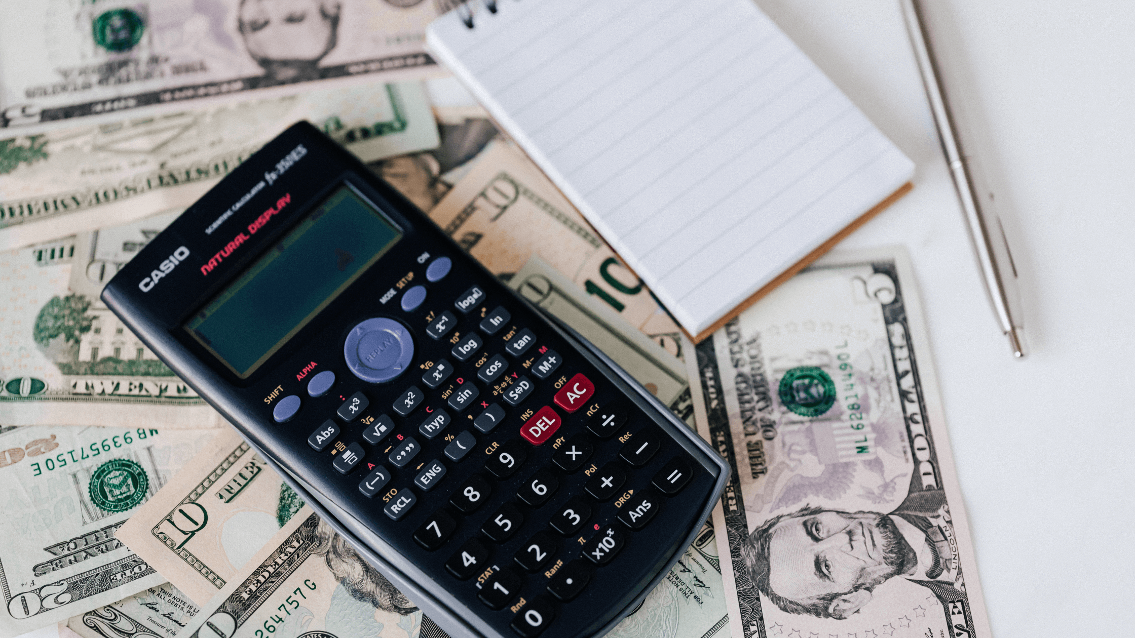 A calculator and a small blank notepad with a pen placed on top of scattered US dollar bills, representing financial management, cash flow analysis, and preparing for tax deadlines.