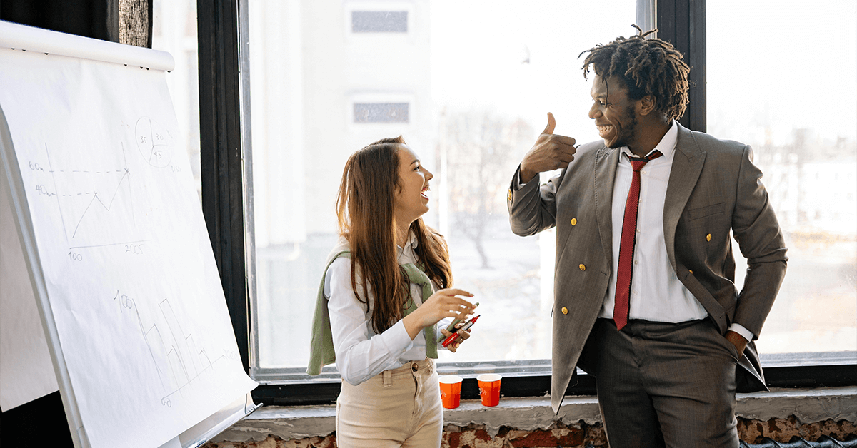 Two business professionals smiling and giving a thumbs-up while standing next to a whiteboard covered in hand-drawn financial charts and graphs, representing successful strategic planning.
